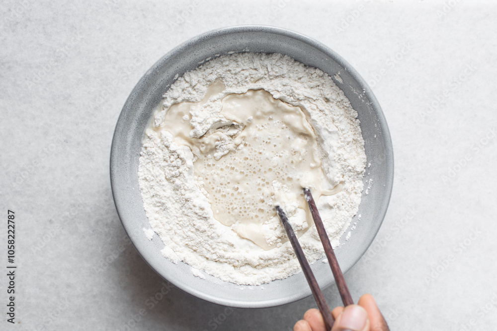 Overhead view of noodle dough being being mixed in a grey bowl, top view of flour and water being mixed with chopsticks to make noodles, process of making hand pulled noodles