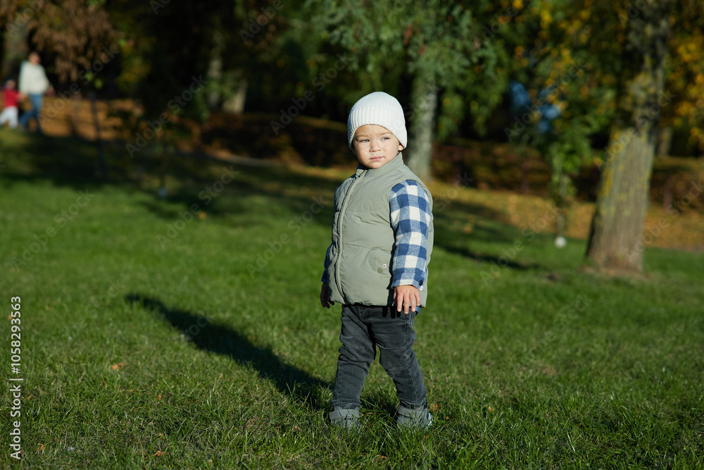A young child stands on green grass in an autumn park, dressed warmly in a beanie, plaid shirt, and vest, looking curiously at the camera. The background is filled with fall foliage. A young child stands on green grass in an autumn park, dressed warmly in a beanie, plaid shirt, and vest, looking curiously at the camera. The background is filled with fall foliage.
