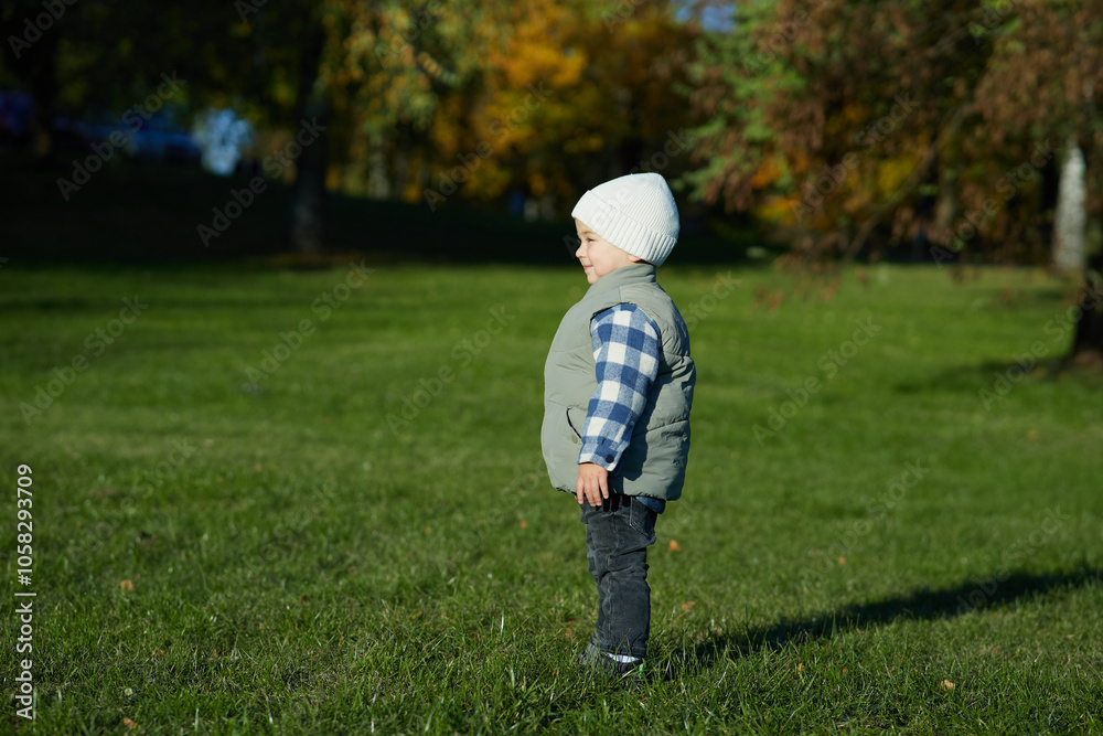 A young child stands on green grass in an autumn park, dressed warmly in a beanie, plaid shirt, and vest, looking curiously at the camera. The background is filled with fall foliage. A young child stands on green grass in an autumn park, dressed warmly in a beanie, plaid shirt, and vest, looking curiously at the camera. The background is filled with fall foliage.