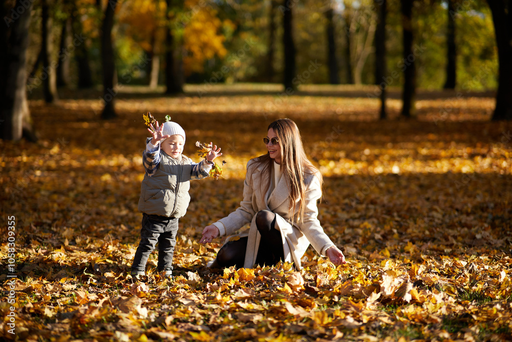 Fototapeta premium A mother and her young child share a tender moment in a park surrounded by autumn leaves. The woman crouches down to her child’s level, creating a warm, intimate scene in golden fall light.