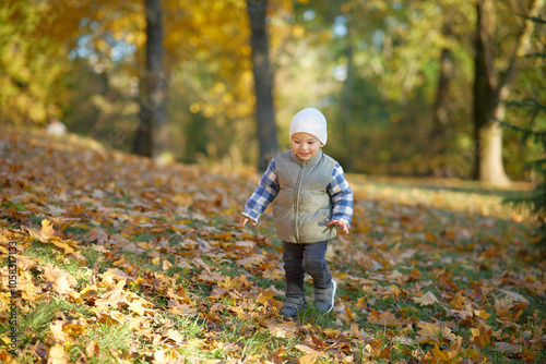 A joyful toddler wearing a cozy hat and vest walks through a park filled with colorful autumn leaves. The child smiles brightly, enjoying the fall season and nature around.