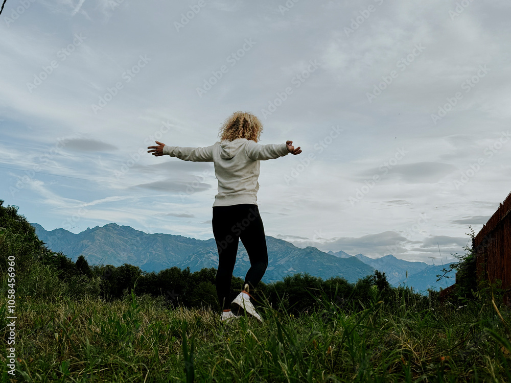 Woman with open arms walks through a green meadow under a blue sky with mountain scenery in the background, expressing freedom, happiness, and nature's beauty. A peaceful outdoor moment Woman with open arms walks through a green meadow under a blue sky with mountain scenery in the background, expressing freedom, happiness, and nature's beauty. A peaceful outdoor moment