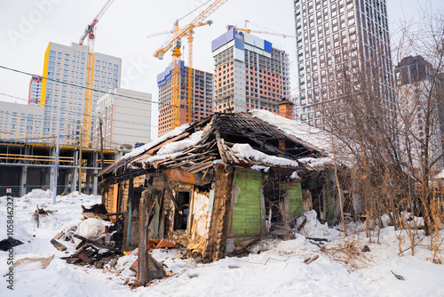 Fotografía A dilapidated wooden house stands in a snowy landscape, overshadowed by towering cranes and modern buildings under construction in a bustling city