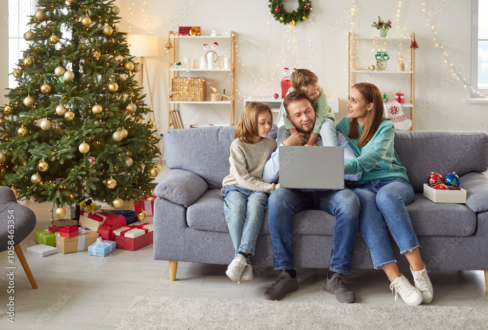 Happy young smiling family with two kids sitting on sofa using laptop together in festive living room with decorated Christmas tree enjoying winter holidays. New Year celebration concept. Happy young smiling family with two kids sitting on sofa using laptop together in festive living room with decorated Christmas tree enjoying winter holidays. New Year celebration concept.