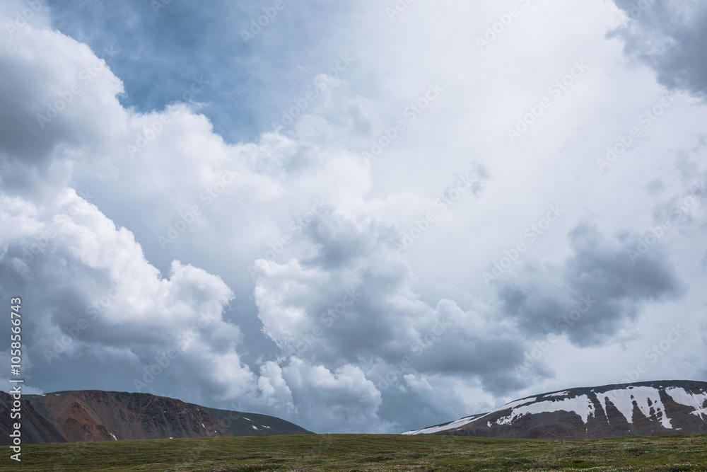 Dramatic minimalist alpine landscape with grassy hill against rocky ridge and snow mountain range under gray cloudy sky. Dark atmospheric view to green hillside in high mountains in changeable weather