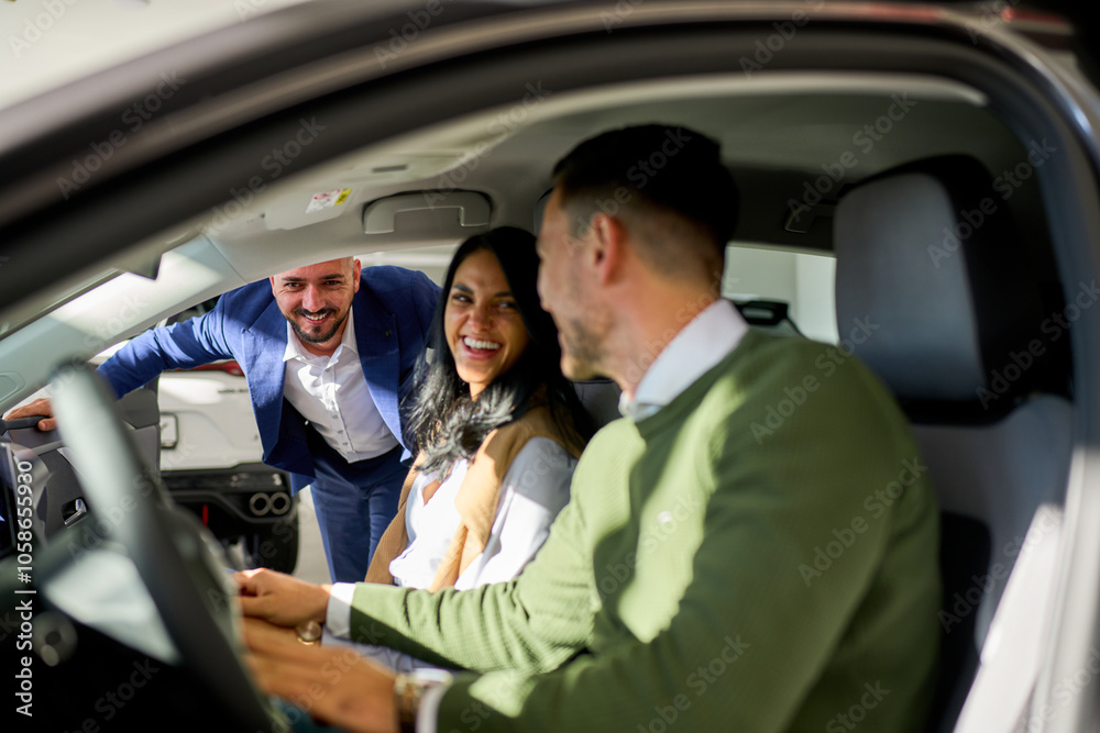 In a car, front seats, a smiling couple faces each other, talking, while a cheerful salesman stands outside, visible through the open window. In a car, front seats, a smiling couple faces each other, talking, while a cheerful salesman stands outside, visible through the open window.