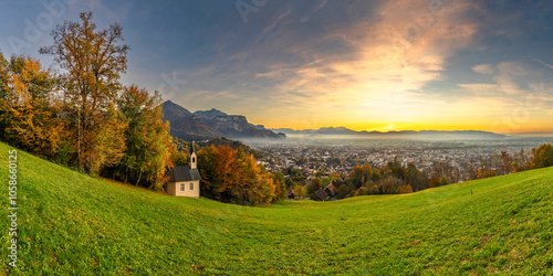 Panorama Sunset in the Rhine Valley, with a tree and a chapel,blue sky over the city of Dornbirn, meadow and fields. with Swiss mountains in the background. afterglow, interesting colored veil clouds