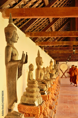 A couple of buddhist monks are looking at the buddha statues, sculptures that are lined up in the cloister, gallery walk, wall structure around the Pha That Luang Stupa, Vientiane, Laos photo