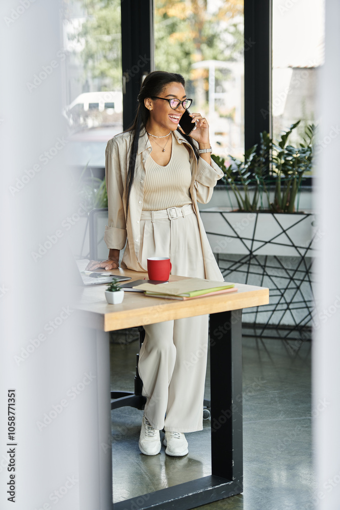 A stylish young woman stands by her desk, chatting on the phone while enjoying her workspace. A stylish young woman stands by her desk, chatting on the phone while enjoying her workspace.