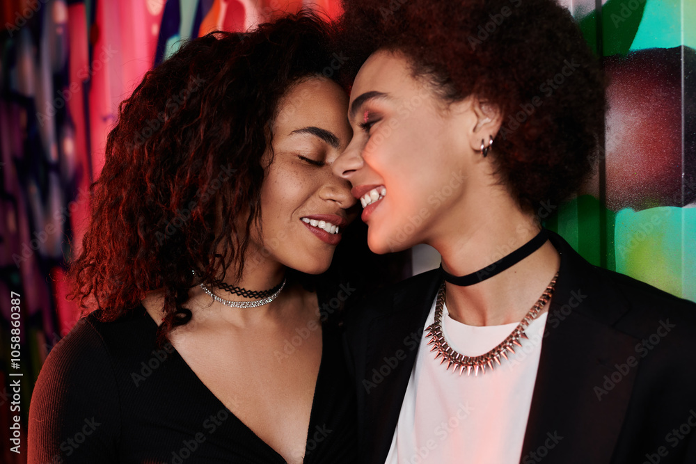 Two women lean close together, smiling affectionately against a colorful wall in the evening. Two women lean close together, smiling affectionately against a colorful wall in the evening.