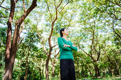 Young Asian woman sits under a shady tree in a park, enjoying her free time in nature. She breathes deeply, refreshing herself, surrounded by leaves and the tranquility of greenery.