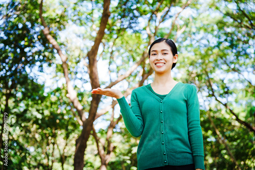 Young Asian woman sits under a shady tree in a park, enjoying her free time in nature. She breathes deeply, refreshing herself, surrounded by leaves and the tranquility of greenery.