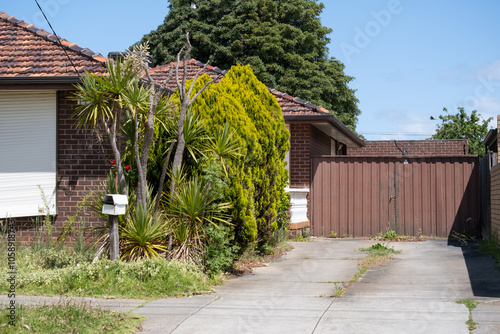 Traditional suburban brick house featuring an overgrown garden and a weedy driveway—weathering suburban residential property with signs of neglect in the front yard in an Australian neighborhood.