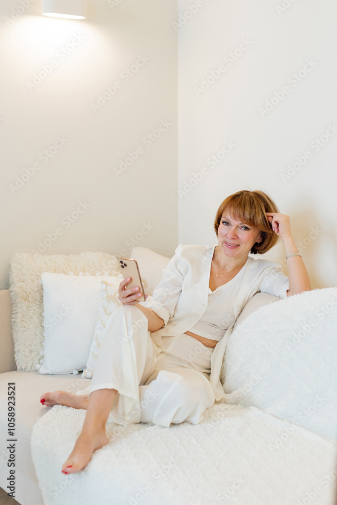 Adult beautiful woman relax siting on sofa at home and using cellphone texting chatting with friend. Happy young woman on couch holding smartphone in her hands, shoping online or check social media. Adult beautiful woman relax siting on sofa at home and using cellphone texting chatting with friend. Happy young woman on couch holding smartphone in her hands, shoping online or check social media.