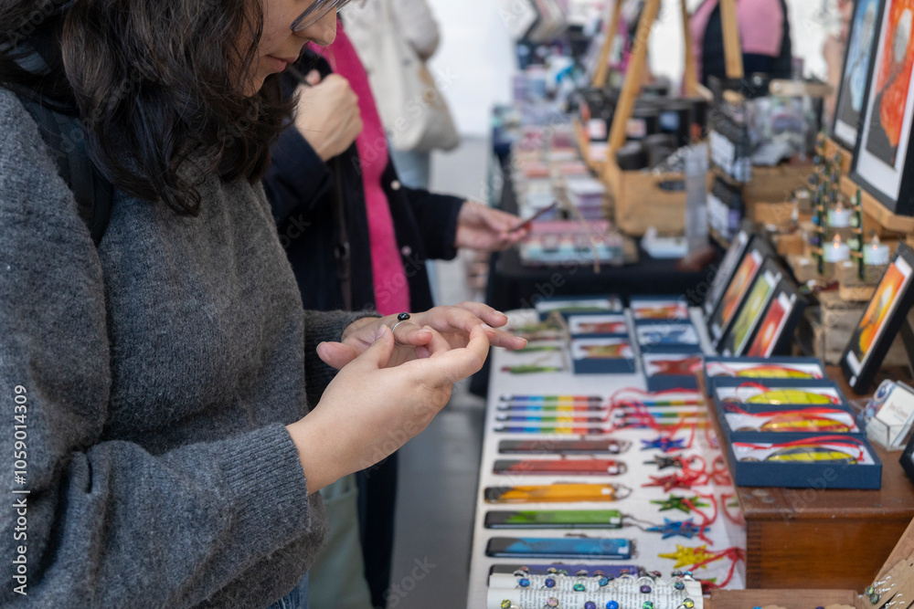 Woman in a gray knit sweater examines items at a bustling craft market. Handmade goods fill the tables in front of her, with other shoppers in the softly blurred background. Woman in a gray knit sweater examines items at a bustling craft market. Handmade goods fill the tables in front of her, with other shoppers in the softly blurred background.