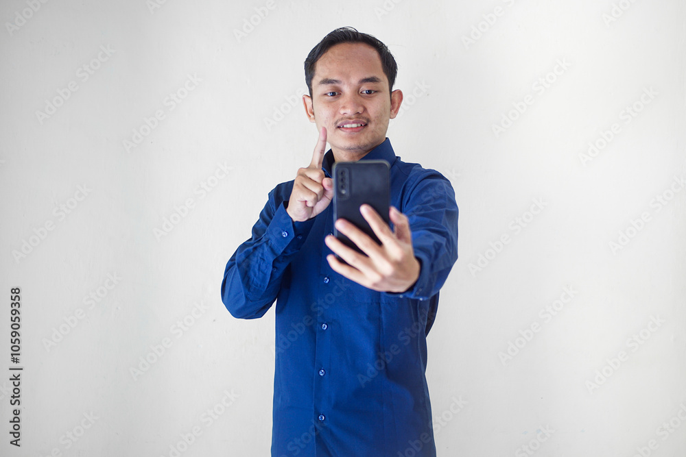 Smiling Young Asian businessman in blue shirt showing finger with one hand use smartphone on white background. Perfect for technology themes, social media interactions and communication gestures Smiling Young Asian businessman in blue shirt showing finger with one hand use smartphone on white background. Perfect for technology themes, social media interactions and communication gestures