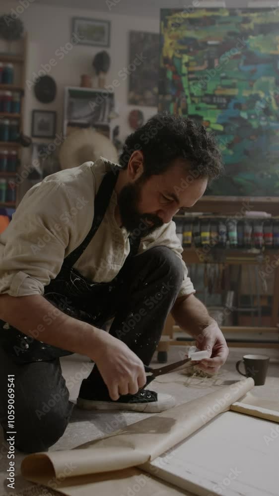 Full vertical shot of mature Caucasian male artist sitting on floor in studio, wrapping finished painting in brown craft paper, fixing with adhesive tape, while packing artwork for sending to buyer Full vertical shot of mature Caucasian male artist sitting on floor in studio, wrapping finished painting in brown craft paper, fixing with adhesive tape, while packing artwork for sending to buyer
