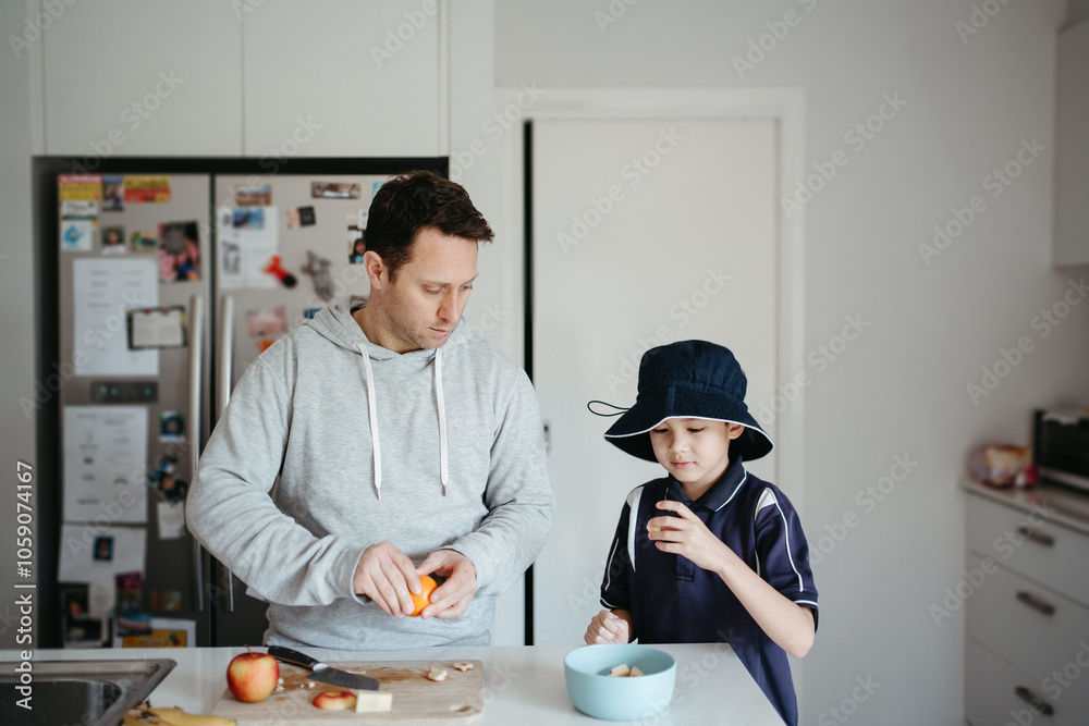 man cutting fruits on the kitchen counter with young boy beside him getting ready for school