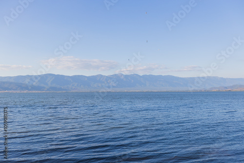 Tranquil Scene Of Doirani Lake With Its Calm Waters Stretching To The Horizon, Framed By Distant Mountain Ranges And A Clear, Blue Sky