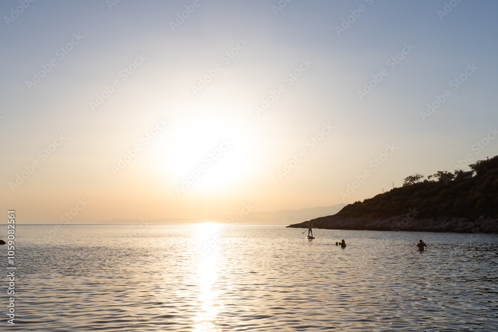 A peaceful summer sunset over calm waters with silhouettes of paddleboarders and swimmers in Greece. A peaceful summer sunset over calm waters with silhouettes of paddleboarders and swimmers in Greece.