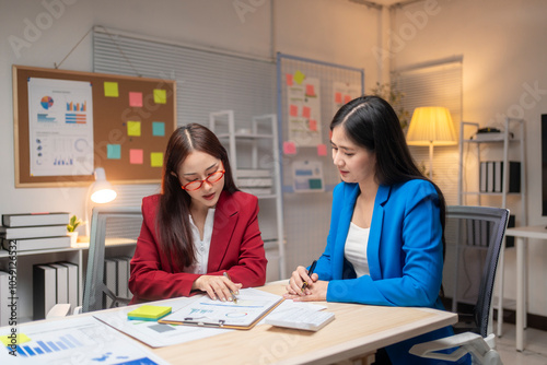 Two women in suits are sitting at a desk with a white board behind them