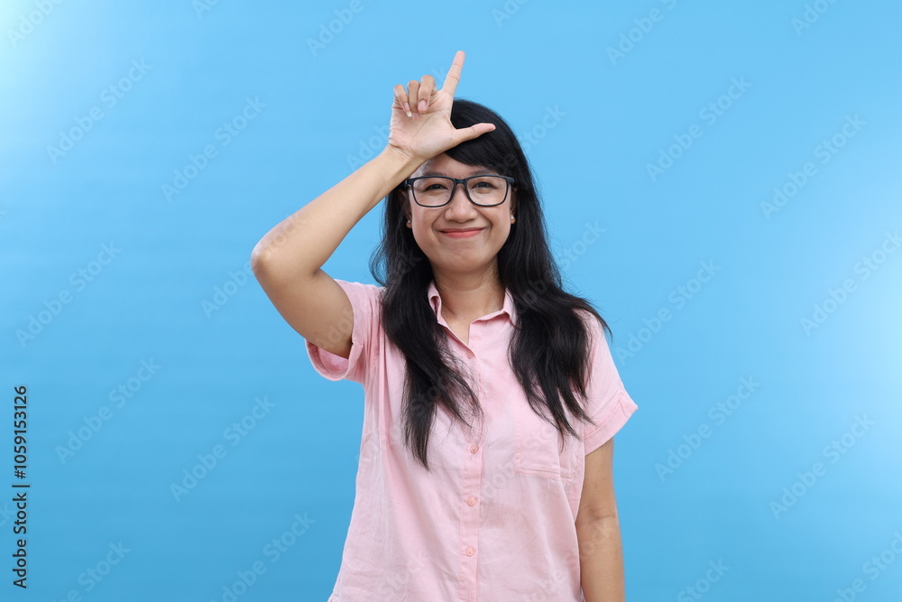 Smiling Asian girl makes loser gesture over forehead, lost shape L ...