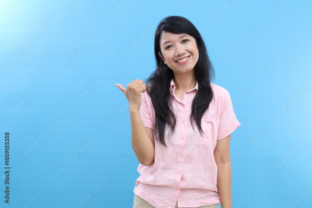 Young cheerful Asian female points away with thumb, happy face expression, demonstrates copy space for advertisement Young cheerful Asian female points away with thumb, happy face expression, demonstrates copy space for advertisement