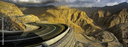 Fotografie Winding mountain road with dramatic shadows and rugged terrain under a cloudy sk
