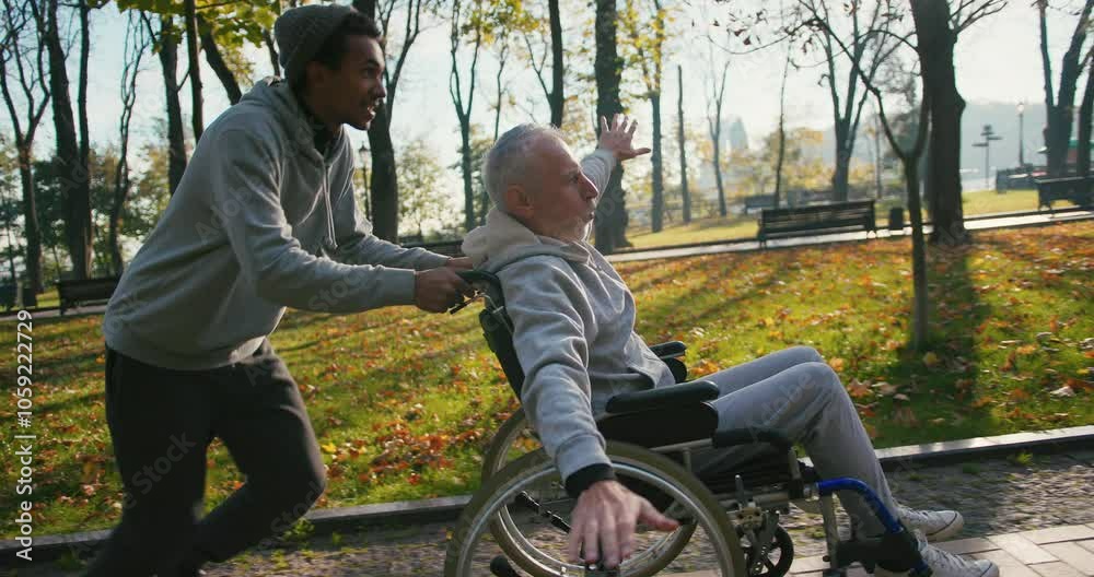 Hispanic man taking care of old disabled friend in wheelchair. Friends running, having fun together in autumn park. Healthcare, disability, mobility, friendship