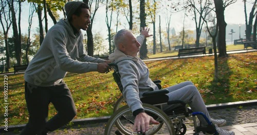 Hispanic man taking care of old disabled friend in wheelchair. Friends running, having fun together in autumn park. Healthcare, disability, mobility, friendship