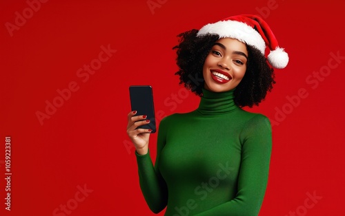 Une femme afro-américaine magnifique portant un col roulé vert et un bonnet de Père Noël tenant un smartphone dans la main, sur fond rouge, image avec espace pour texte. photo