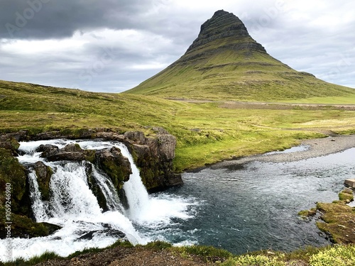 Iceland’s iconic Kirkjufell mountain with a scenic waterfall in the foreground. The lush green landscape and overcast sky create a serene, atmospheric view, ideal for nature and travel themes.