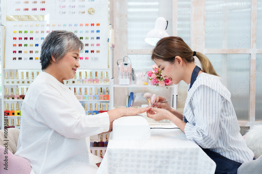 Obraz premium Young female nail technician carefully manicures hand of senior Asian woman in bright nail salon. Technician in striped shirt intently on client’s nails amid vibrant nail polish display, and relaxing