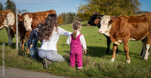 Mutter mit zwei Kindern (2 Jahre alt) vor Kuhweide, Ausflug mit Zwillingen, Junge und Mädchen in Latzhosen, vor Kuhherde in Bayern, blauer Himmel, breites Bildformat,