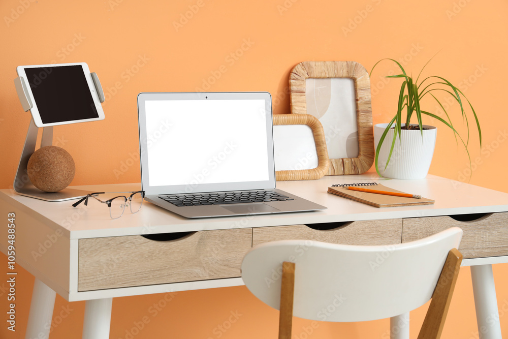 Stylish table with modern laptop and tablet near orange wall in room Stylish table with modern laptop and tablet near orange wall in room