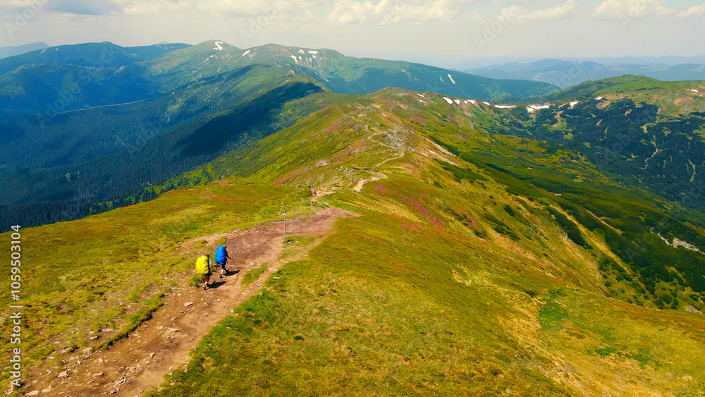 Fototapeta premium Hikers on a mountain ridge with scenic views of rolling hills. A breathtaking view of two hikers walking along a mountain ridge, surrounded by lush green hills and distant mountain peaks.