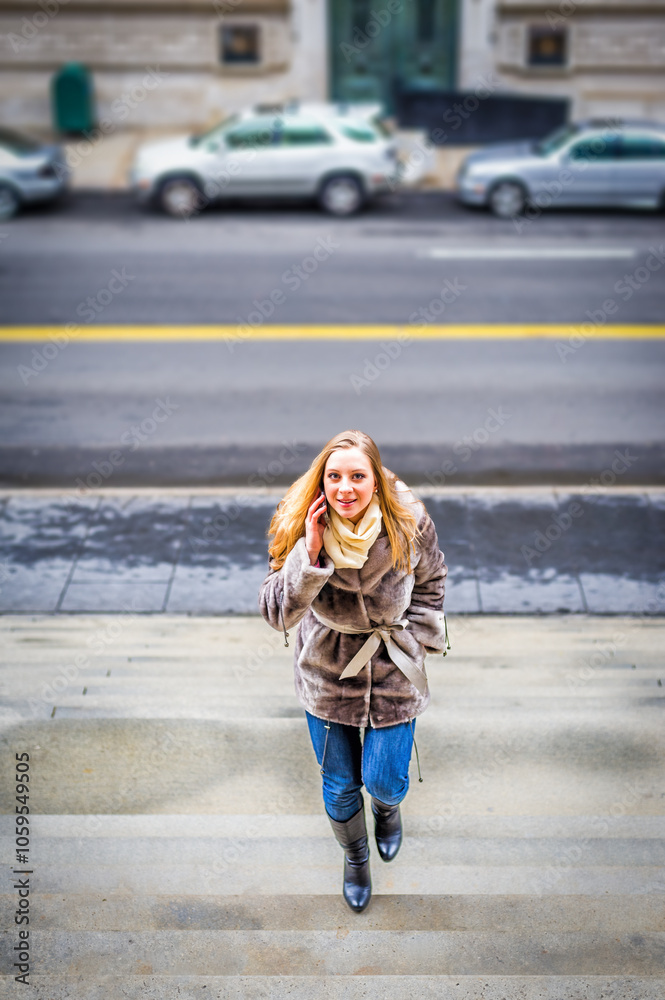 A woman in a fur coat walks up a set of stone steps on a city street. Cars are parked along the road, and she appears to be mid-conversation on a phone.