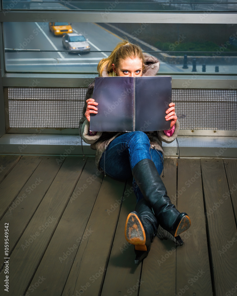 A woman sits on a wooden floor, partially hiding behind a large open book. A window behind reveals a view of a highway with passing vehicles.