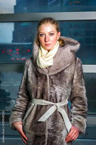 A woman stands indoors wearing a luxurious, fur-lined coat with a hood, complemented by a cream-colored scarf. The background features an urban scene with blurred traffic lights.