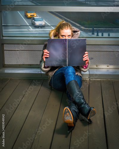 Obraz na płótnie A woman sits on a wooden floor, partially hiding behind a large open book. A window behind reveals a view of a highway with passing vehicles. w salonie
