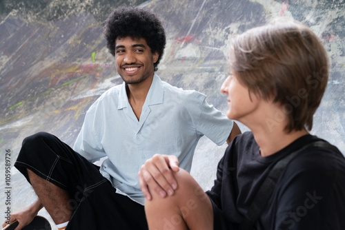 Two young men sitting in a skatepark, smiling and talking.

