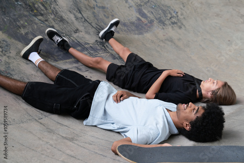  Two young men lying in a skate bowl, smiling with skateboard nearby
