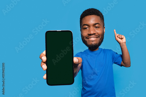 A cheerful young man proudly displays a smartphone against a vibrant blue backdrop while celebrating a moment of joy photo