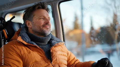 A man in a bright orange winter jacket, seated inside a vehicle, enjoys the snowy view beyond the window, embodying adventure and the spirit of exploration.