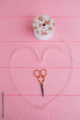 Lace ribbon, elegantly wrapped in the shape of a heart on a pink background, and in the middle lie metal scissors that add charm to DIY projects and crafts photo