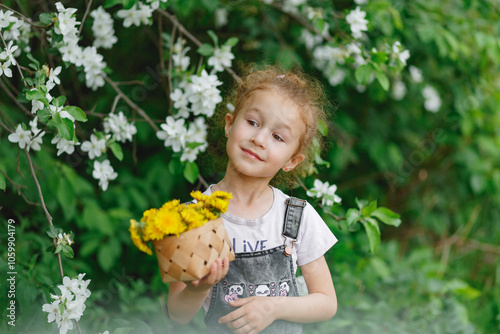 Little curly girl sniffing flowers of blossoming apple tree with dandellions in her hands. garden with flowering trees photo