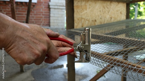 male hands holding red handled construction pliers and clamping metal mesh to make a rim of the base of a quail cage, working with a clamp in the process of creating an agricultural cage photo