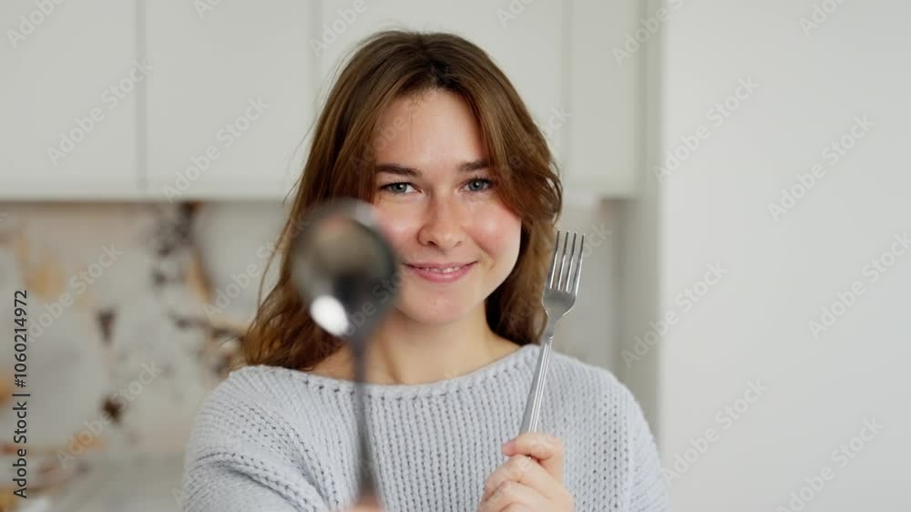 Young woman smiles while holding a fork and spoon in a sunny kitchen. Radiating joy and playfulness, she prepares a meal with enthusiasm and delight