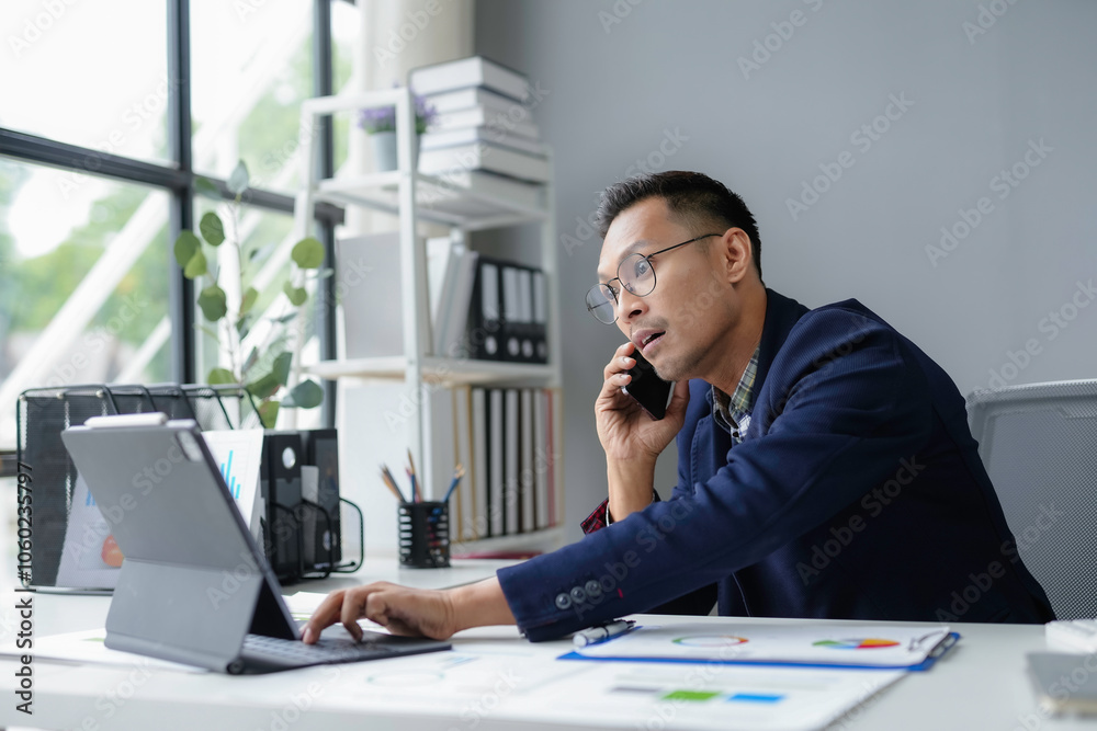 Naklejka premium Businessman is multitasking, talking on his phone while working on his tablet in his office