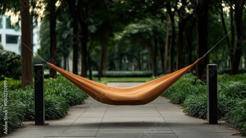 Orange hammock suspended between two poles in a tree-lined park pathway surrounded by green foliage.